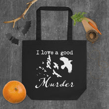 Load image into Gallery viewer, A black tote bag with a white graphic of a murder of crows with the words, "I love a good Murder" laying flat on a gray surface with halloween decorations around it.